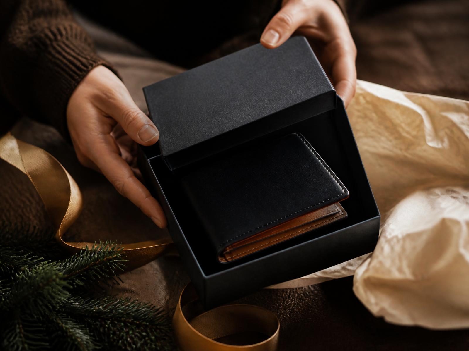 Hands presenting a black leather wallet with tan interior inside a dark gift box